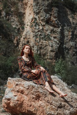 Güçlü iradeli, güçlü, kızıl saçlı, etnik elbiseli bir kadın büyük bir taşın yanında. Yenilmezliğin sembolü. Kariyer