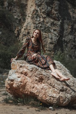 Güçlü iradeli, güçlü, kızıl saçlı, etnik elbiseli bir kadın büyük bir taşın yanında. Yenilmezliğin sembolü. Kariyer