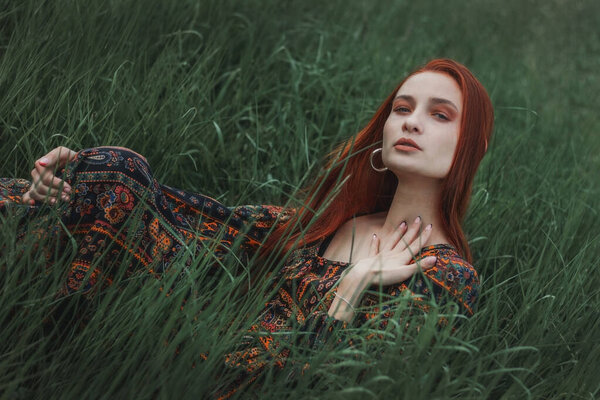 Portrait of a red-haired girl dressed in an ethnic dress. Green grass. Mountain. Merging with nature. Concept