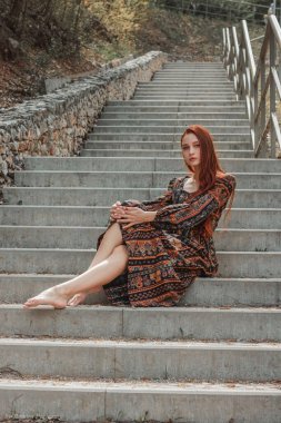 Uzun boylu, kızıl saçlı, kendine güvenen, etnik elbiseli bir kız merdivenlerde oturuyor. Kızıl saçlar rüzgârda gelişir. Kavram. İradenin ve boyun eğmezliğin sembolü.