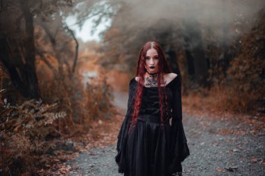 Gotik kadın, sonbahar ormanında kızıl saçlı cadı, siyah elbiseli sis, Cadılar Bayramı. Mistik atmosfer
