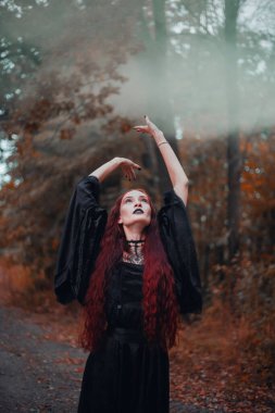 Gotik kadın, sonbahar ormanında kızıl saçlı cadı, siyah elbiseli sis, Cadılar Bayramı. Mistik atmosfer