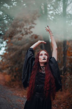 Gotik kadın, sonbahar ormanında kızıl saçlı cadı, siyah elbiseli sis, Cadılar Bayramı. Mistik atmosfer