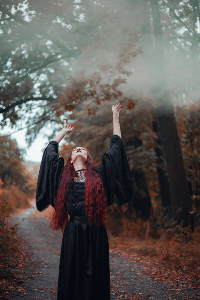 Goth woman, red-haired witch in autumn forest in fog in black dress, Halloween. Mystical atmosphere