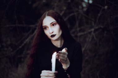 Siyah kostümlü cadı karanlık bir ormanda mum tutuyor. Gizemli atmosfer, ayin, cadılar bayramı.