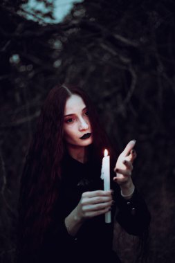 Siyah kostümlü cadı karanlık bir ormanda mum tutuyor. Gizemli atmosfer, ayin, cadılar bayramı.