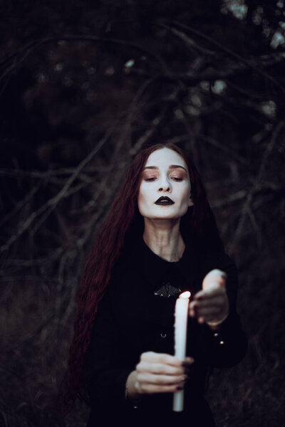 Ведьма в черном костюме держит свечу в темном лесу, таинственная атмосфера, ритуал, Хэллоуин