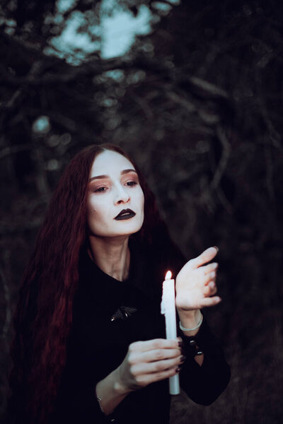 Ведьма в черном костюме держит свечу в темном лесу, таинственная атмосфера, ритуал, Хэллоуин