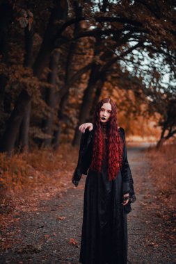 Cadı gibi giyinmiş siyah elbiseli kızıl saçlı kadın portresi, gotik. Cadılar Bayramı, sonbahar ormanı
