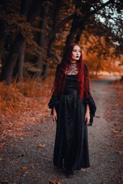 Cadı gibi giyinmiş siyah elbiseli kızıl saçlı kadın portresi, gotik. Cadılar Bayramı, sonbahar ormanı