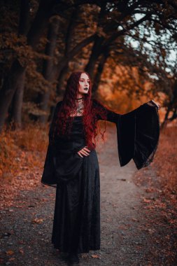 Cadı gibi giyinmiş siyah elbiseli kızıl saçlı kadın portresi, gotik. Cadılar Bayramı, sonbahar ormanı