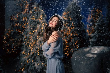 Kadın kış ormanında kar yağışı ile gülümsüyor. Evergreen ağaçları ve tatil ışıkları, Noel ve yeni yıl kutlamaları için şenlikli atmosfer.