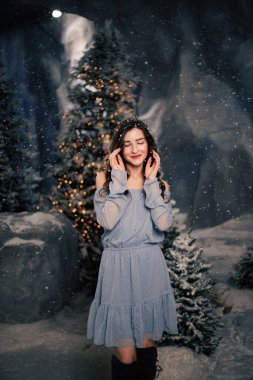 Kadın kış ormanında kar yağışı ile gülümsüyor. Evergreen ağaçları ve tatil ışıkları, Noel ve yeni yıl kutlamaları için şenlikli atmosfer.