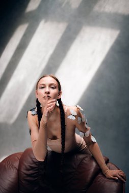 Uzun örgülü bir kadın, gün boyu güneşli bir odada teninde kelebekler olan bir sandalyeye poz verir.