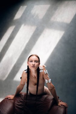 Uzun örgülü bir kadın, gün boyu güneşli bir odada teninde kelebekler olan bir sandalyeye poz verir.