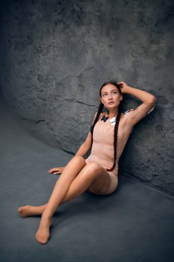 Saçları örgülü ve omuzlarında kelebekler olan genç bir kadın yumuşak ışıklandırmada dokulu bir duvara karşı duruyor.