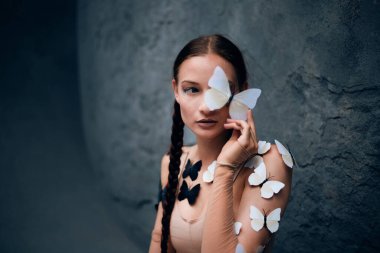 Kadın, gündüz vakti açık havada bir duvarda kelebekler dolaşırken bir kelebeği yüzüne yakın tutuyor.