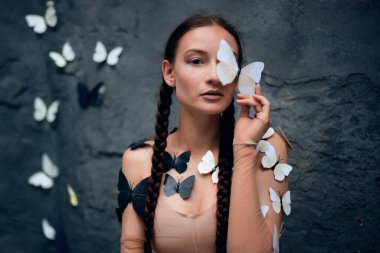 Kadın, gündüz vakti açık havada bir duvarda kelebekler dolaşırken bir kelebeği yüzüne yakın tutuyor.