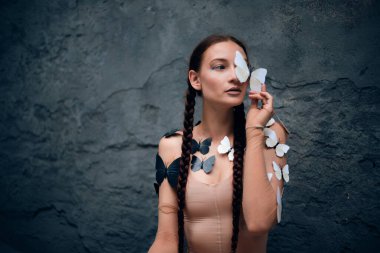 Kadın, gündüz vakti açık havada bir duvarda kelebekler dolaşırken bir kelebeği yüzüne yakın tutuyor.