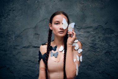 Kadın, gündüz vakti açık havada bir duvarda kelebekler dolaşırken bir kelebeği yüzüne yakın tutuyor.