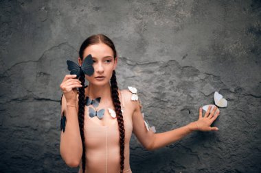 Kadın, gündüz vakti açık havada bir duvarda kelebekler dolaşırken bir kelebeği yüzüne yakın tutuyor.