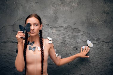 Kadın, gündüz vakti açık havada bir duvarda kelebekler dolaşırken bir kelebeği yüzüne yakın tutuyor.