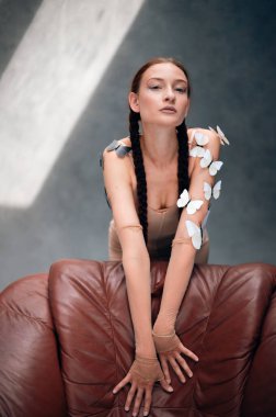 Uzun örgülü bir kadın, gün boyu güneşli bir odada teninde kelebekler olan bir sandalyeye poz verir.