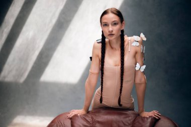 Uzun örgülü bir kadın, gün boyu güneşli bir odada teninde kelebekler olan bir sandalyeye poz verir.