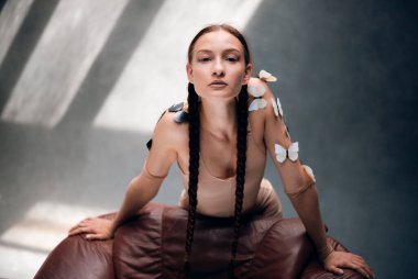 Uzun örgülü bir kadın, gün boyu güneşli bir odada teninde kelebekler olan bir sandalyeye poz verir.