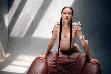 Uzun örgülü bir kadın, gün boyu güneşli bir odada teninde kelebekler olan bir sandalyeye poz verir.