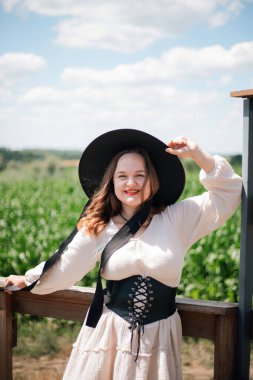 Doğanın tadını çıkaran genç bir kadın, güneşli bir yaz gününde mısır tarlasının yakınında şapkasıyla gülümsüyor.