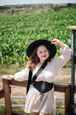 Doğanın tadını çıkaran genç bir kadın, güneşli bir yaz gününde mısır tarlasının yakınında şapkasıyla gülümsüyor.