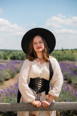 Beyaz elbiseli ve siyah şapkalı bir kadın güneşli bir yaz gününde lavanta tarlasında poz veriyor.