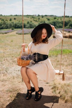 Beyaz elbiseli ve siyah şapkalı bir kadın, elinde saman balyalarıyla güneşli kırsal bir ortamda sepeti tutarken sallanıyor.