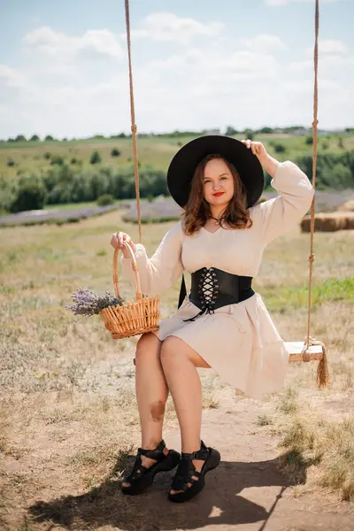 Beyaz elbiseli ve siyah şapkalı bir kadın, elinde saman balyalarıyla güneşli kırsal bir ortamda sepeti tutarken sallanıyor.