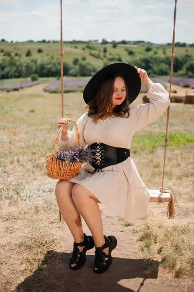 Beyaz elbiseli ve siyah şapkalı bir kadın, elinde saman balyalarıyla güneşli kırsal bir ortamda sepeti tutarken sallanıyor.