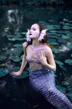 Deniz kızı, akşam ışığında yeşil bitkiler ve suyla çevrili bir göldeki bir nilüferde poz verir.