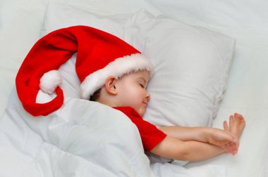 Küçük çocuk Noel Baba şapkalı beyaz çarşafların üzerinde uyuyor..
