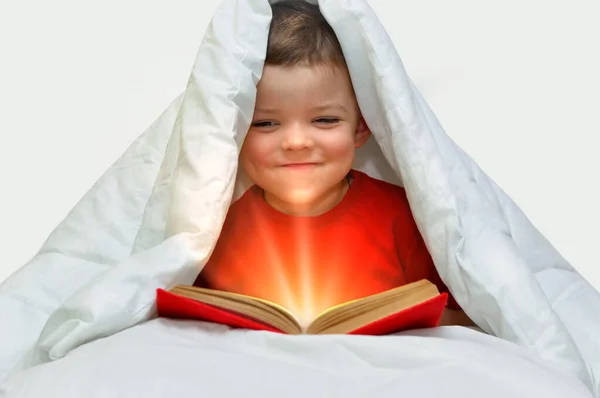 A little boy in a red t-shirt sits under a blanket with a book that glows inside