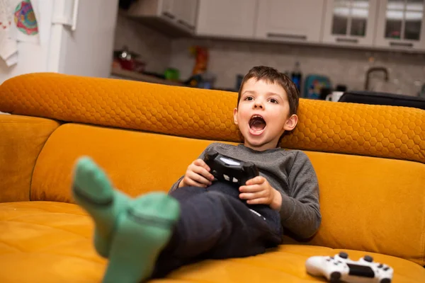 stock image Cute 5 year old boy playing video game console while sitting on orange couch at home.