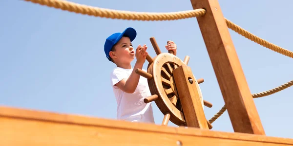 A little boy in light clothes stands and holds a wooden steering wheel in an eco-friendly wooden playground in Europe.