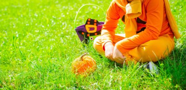 Turuncu balkabağı kostümü giymiş, elinde bir çanta şeker ve balkabağıyla cadılar bayramında yeşil çimlerin üzerinde oturan bir çocuğun geniş fotoğrafı.