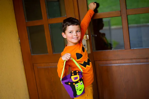 A seven-year-old boy dressed as a pumpkin and carrying a candy bag knocks on the door and smiles.