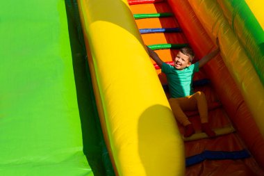 Parlak renkli elbiseli küçük bir çocuk güneşli bir yaz gününde çok renkli bir kaydırağın merdivenlerinde oturur ve gülümser.