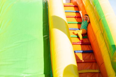 Parlak renkli elbiseli küçük bir çocuk çok güneşli bir yaz gününde parktaki renkli bir kaydırağın merdivenlerinde oturur, gülümser ve el sallar. Güneşten kaçarken elleriyle gözlerini kapatıyor.