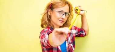 Kırmızı kareli gömlekli, güvenlik gözlüğü takan güzel bir kadın elinde bir mezura tutuyor.