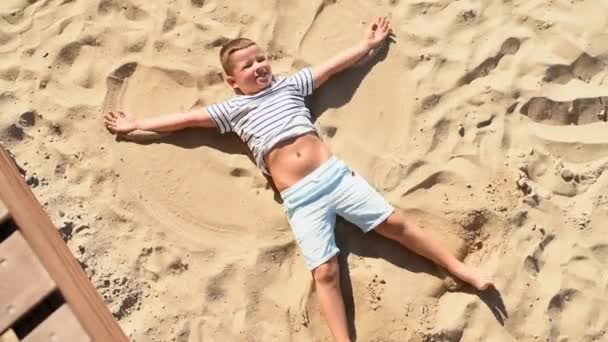 白いストライプTシャツと青い夏のショートパンツの幸せな子供の少年は砂の上にあり 手と足で天使を作ります — ストック動画