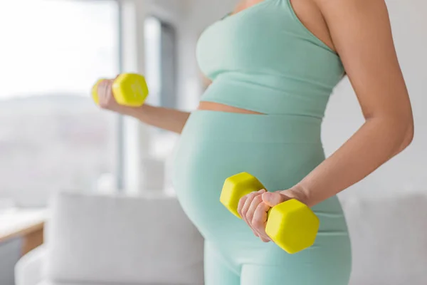 Pregnant woman prenatal training at home doing fitness strength training exercise with dumbbell weights. Body workout arm curls closeup of belly and arms. Easy workout in second and third timester.