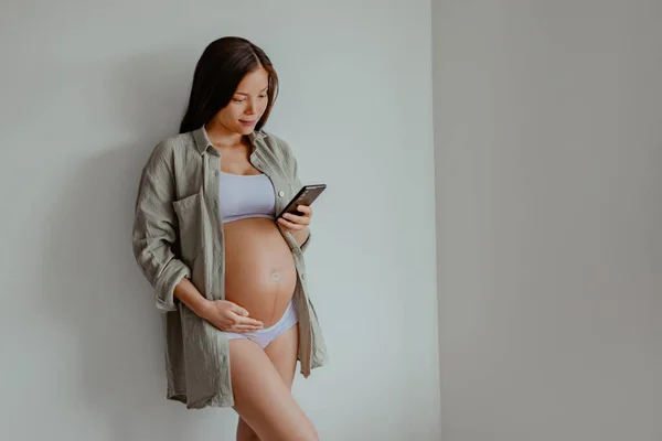 Pregnant woman using phone app for online shopping maternity clothing or reading about labor and birth wearing underwear and casual cotton shirt at home. Relaxing holding expecting belly for health.