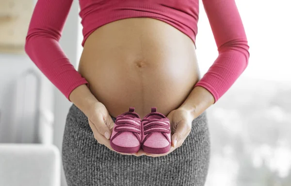 怀孕了怀孕妇女期待女婴抱着粉色红鞋的性别透露 等妈妈表现出宝宝肚子肿大 婴儿服装 — 图库照片
